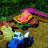 Muoviset kuorma-auto ja traktori, hiekkakakkumuotteja, pikkulapio ja muita leluja hiekkalaatikolla.