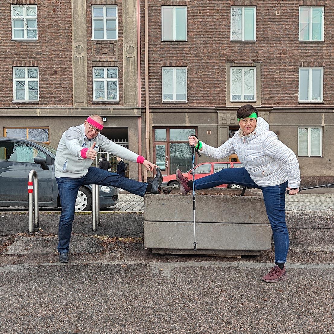 Jarkko Eloranta ja Katja Syvärinen venyttelevät betonisen ulkokukkalaatikon vieressä hikipannat päässään, Katjalla lisäksi kävelysauvat.