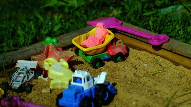 Muoviset kuorma-auto ja traktori, hiekkakakkumuotteja, pikkulapio ja muita leluja hiekkalaatikolla.