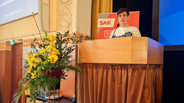 Katja Syvärinen Paasitornin puhujapöntössä pitämässä puhetta, vieressä kaunis kukka-asetelma.