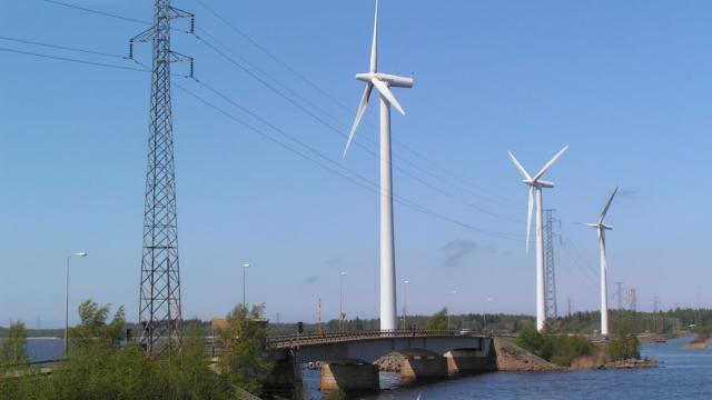 Voimalinjan pylväs ja kolme tuulivoimalaa järven ylittävän sillan ja penkereen vieressä.