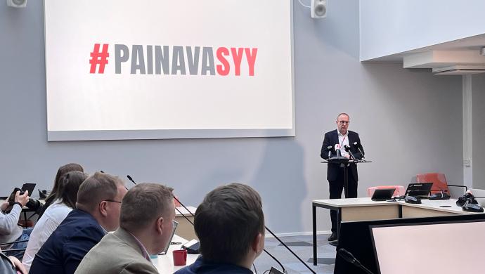 Jarkko Eloranta puhuu mediatilaisuudessa toimittajille, taustalla #PainavaSyy-logo.