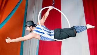 Sirkustaitelija on vatsallaan sirkusteltan katosta vaijerilla roikkuvassa hulavanteessa ja pitää yhdellä kädellä kiinni vanteesta.