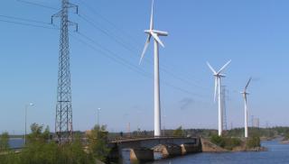 Voimalinjan pylväs ja kolme tuulivoimalaa järven ylittävän sillan ja penkereen vieressä.