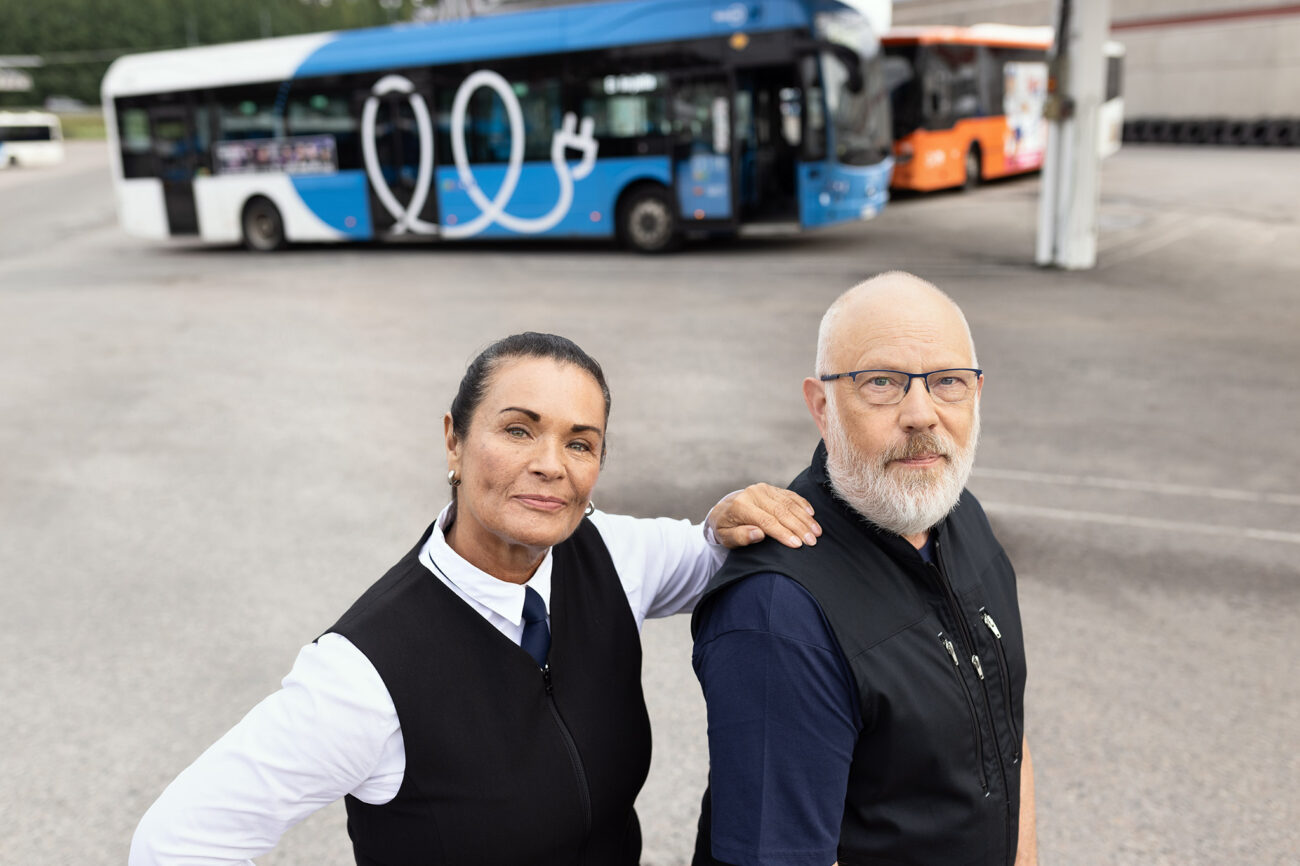 Bussikuski ja siivooja seisovat bussien edessä ja bussikuski pitelee kättään siivoojan olkapäällä.
