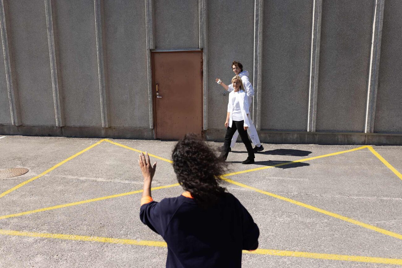 Henkilö seisoo selin kameraan päin ja tervehtii kahta ohikulkevaa työkaveriaan nostamalla käden ylös.