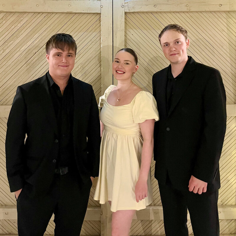 Glow Up Trion kolme muusikkoa seisovat paneeliseinän edessä juhlavasti pukeutuneena.