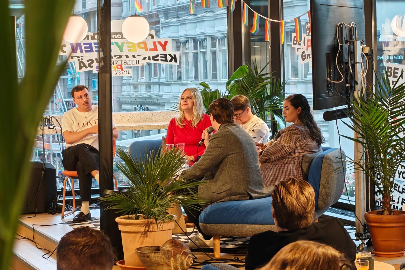 Neljä panelistia ja juontaja istuvat puoliympyrän muotoisesti, taustalla iso ikkuna, jossa Helsinki Pride -tarroja.