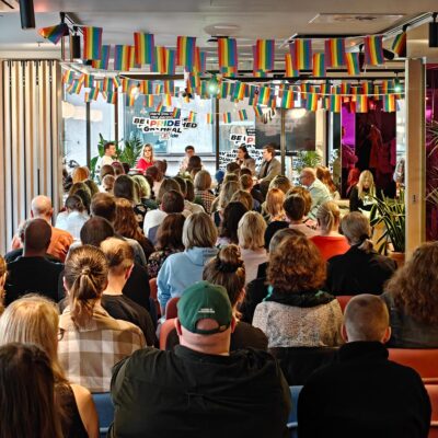 Paljon ihmisiä istumassa ja seuraamassa paneelikeskustelua, katosta roikkuu pieniä Pride-lippuja.