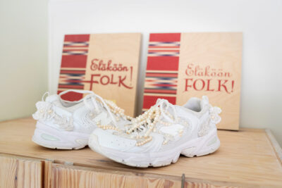 Ett par vita sneakers som är utsmyckade med pärlor står på en trälåda, och i bakgrunden syns två likadana böcker med texten ”Eläköön FOLK!” på pärmen.