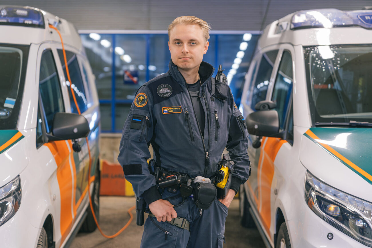 Rajatarkastaja Roope Rasi on pukeutunut rajavartijan työhaalariin ja työkaluvyöhön, seisoo kahden Rajavartiolaitoksen pakettiauton välissä ja katsoo suoraan kameraan.