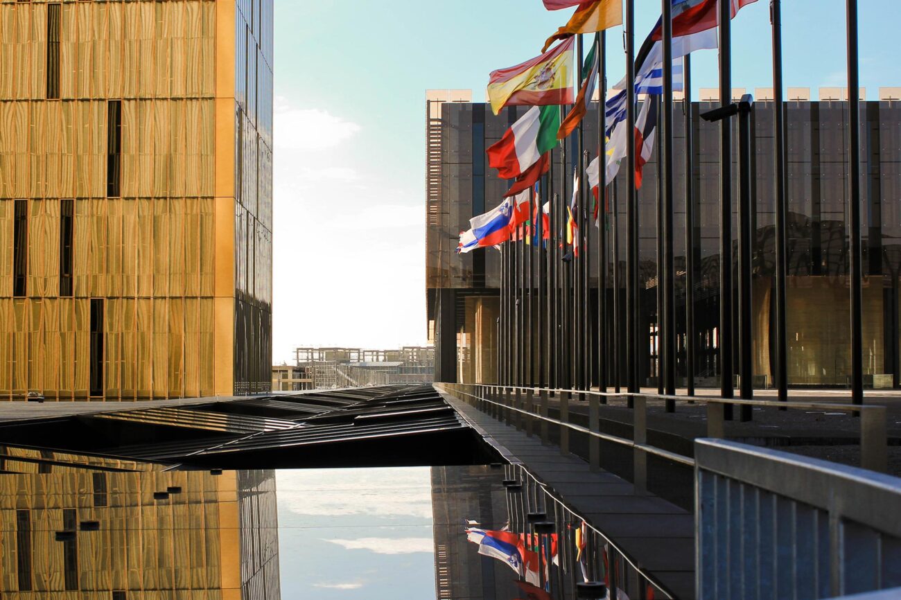 Kaksi modernia rakennusta, joiden välissä on vesiaihe, josta heijastuu toisen rakennuksen edessä oleva lippurivistö.