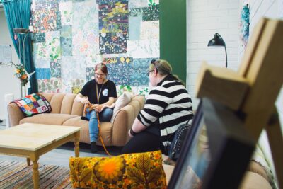 Projektarbetaren Elisa Kivinen och en kund i kulturverkstaden sitter på en soffa, medan Kivinen håller ett färggrant stickat arbete i sina händer.