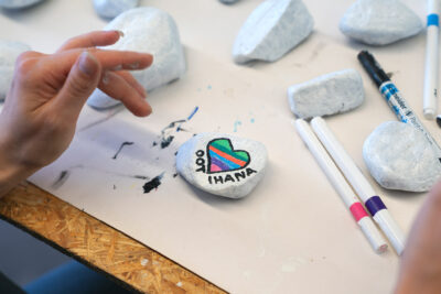 Färgpennor och ljusa små stenar på ett bord. På en av stenarna har någon ritat ett färggrant hjärta och skrivit texten ”Oot ihana” (”Du är underbar”).