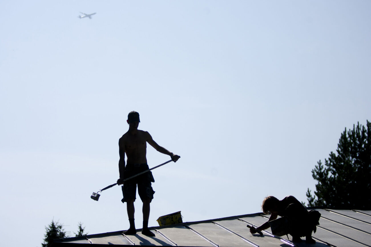 Kaksi kattomaalaria tekee maalaustöitä peltikatolla hellepäivänä.