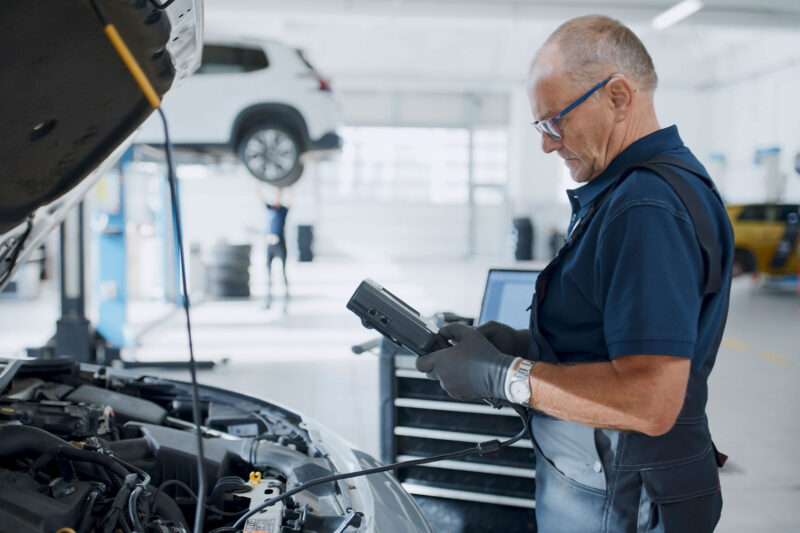 Ikääntynyt mekaanikko tutkii autoa diagnostikkalaitteella.