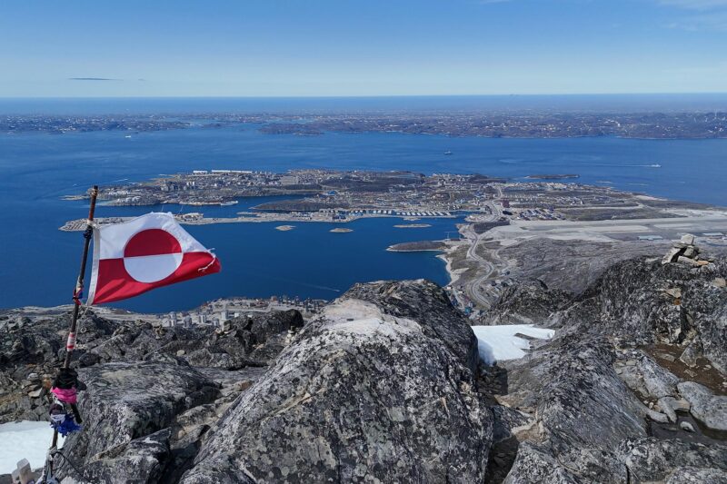 Vuoren kallioisella laella liehuu Grönlannin lippu ja sieltä avautuu näkymä kaupunkiin ja merelle.