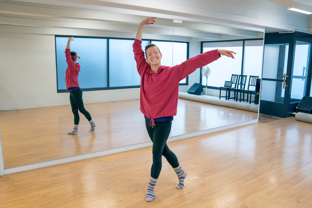 Pau Kettunen står framför spegeln i en danssal i en danspose: med händerna utsträckta uppåt och åt sidan och ena benet bakom den andra.