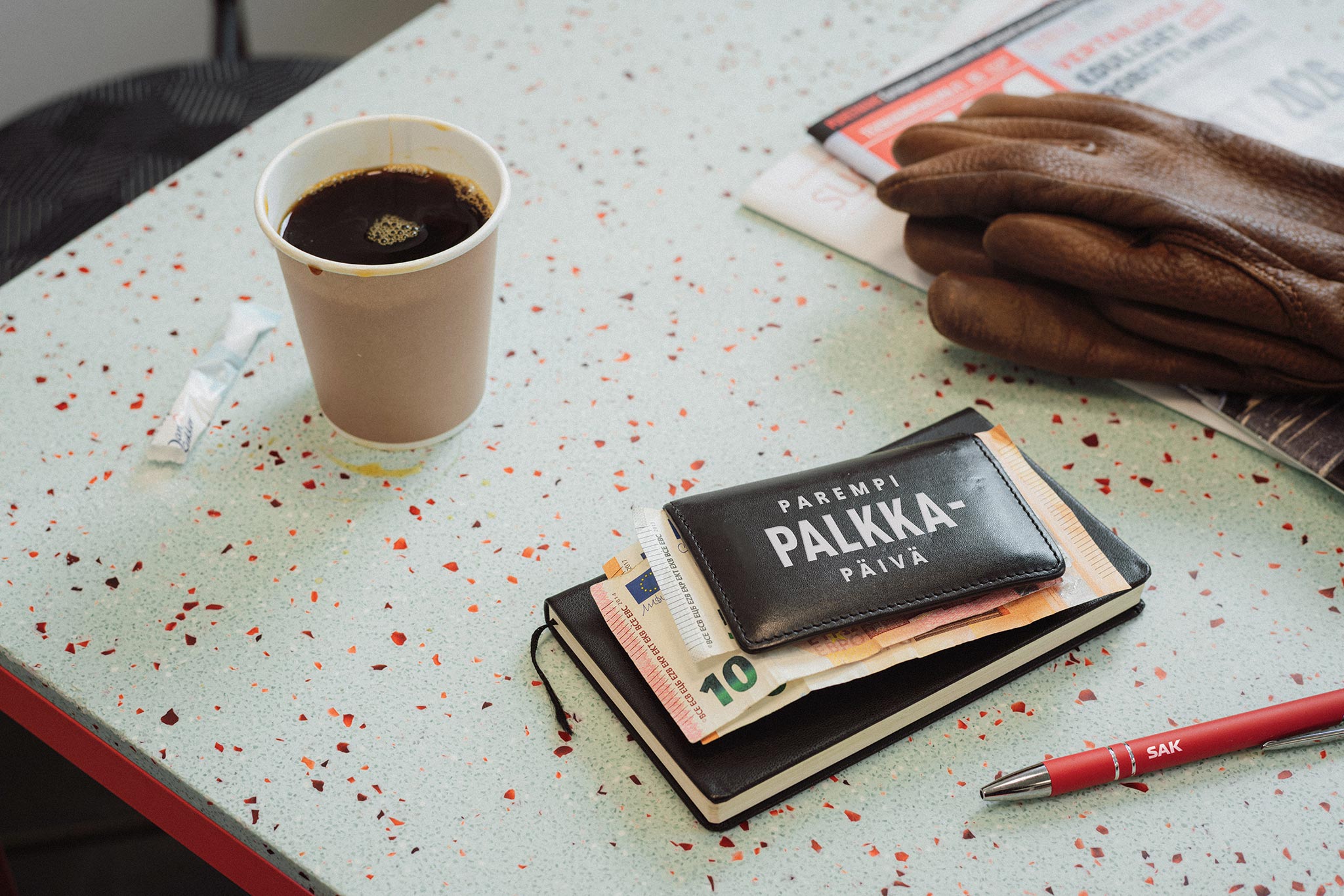 Pöydällä paperinen täysi kahvimuki, kaksi sanomalehteä, ruskeat nahkahansikkaat, kynä ja muistikirja, jonka päällä seteleitä ja lompakko, jonka kannessa lukee "Parempi palkkapäivä".
