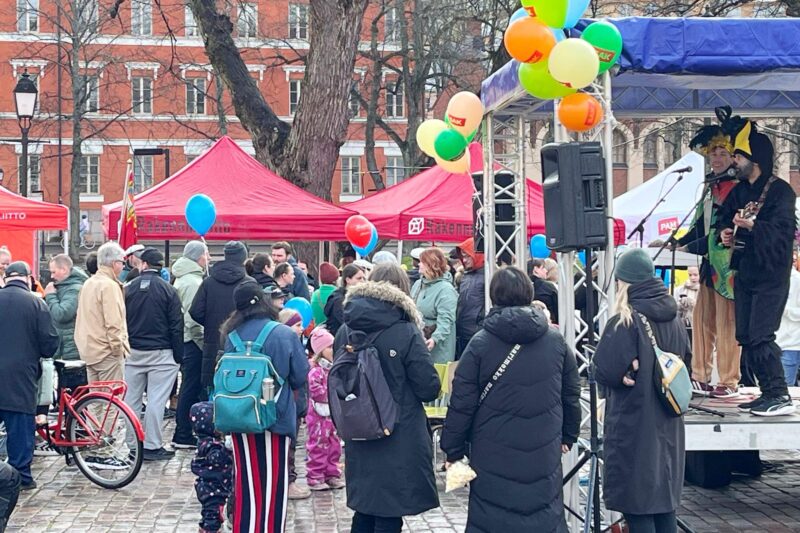 Paljon ihmisiä ulkona esiintymislavan edessä, lavalla kitaristi ja kosketinsoittaja vappuisesti pukeutuneina, lavan katoksessa kiinni paljon värikkäitä ilmapalloja.