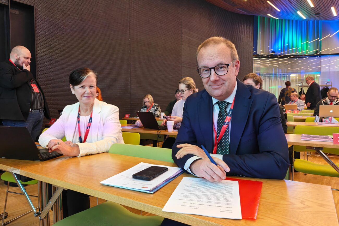 Katja Syvärinen ja Jarkko Eloranta istuvat pöydän ääressä ja katsovat suoraan kameraan, taustalla edustajiston jäseniä istumassa.