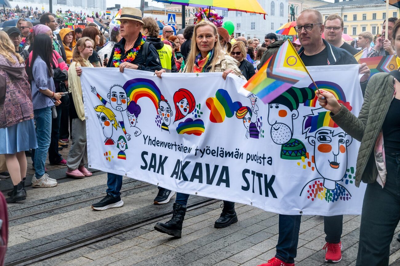 Antti Palola, Maria Löfgren ja Jarkko Eloranta pitelevät värikästä banderollia edessään ja kävelevät kulkueessa.