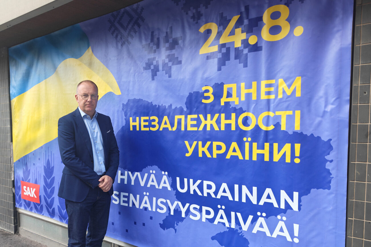 Jarkko Eloranta seisoo Ukraina-aiheisen banderollin edessä, banderollissa Ukrainan lipun värit keltainen ja sininen, päivämäärä 24.8. ja teksti "Hyvää Ukrainan itsenäisyyspäivää!" ukrainaksi ja suomeksi.