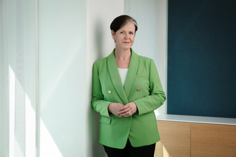Katja Syvärinen on pukeutunut vihreään pikkutakkiin ja nojaa seinään.
